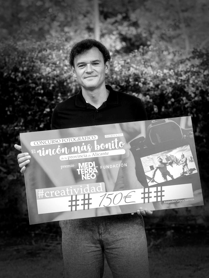 Black and white photo of artist Lejandrier outdoors holding a large poster announcing he is the winner of a photography open call