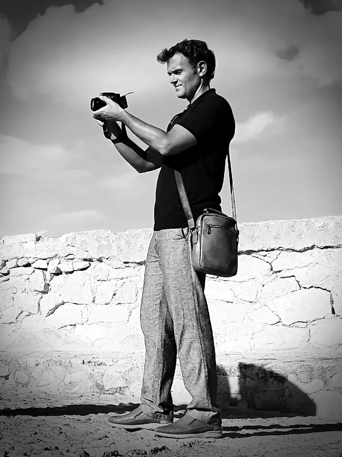 Man taking a photo with a camera against a stone wall and cloudy sky.